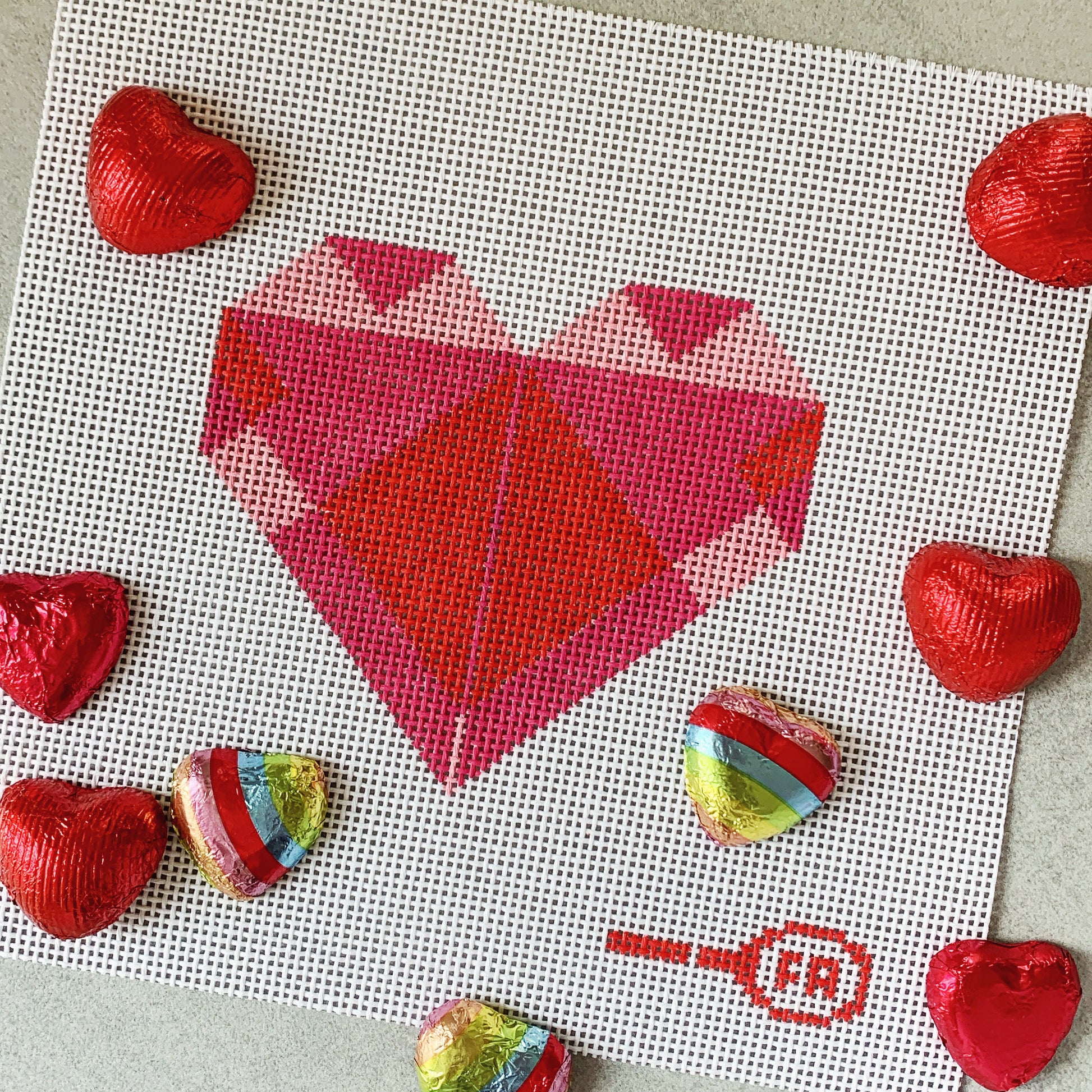 Flat-lay of Origami Heart needlepoint canvas kit—geometric heart motif in shades of red and pink printed on natural 14-mesh canvas with a Frances Abel icon border, styled on a surface scattered with foil-wrapped chocolate hearts