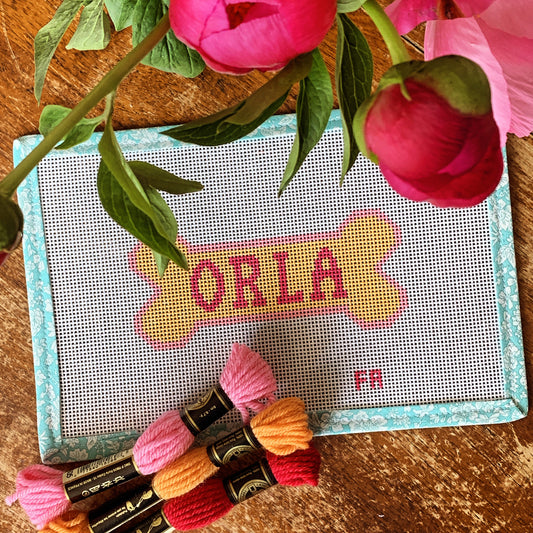 Custom dog bone needlepoint canvas with a yellow bone outlined in red, featuring space for a pet name, hand-painted on white 13 mesh with aqua floral trim.
