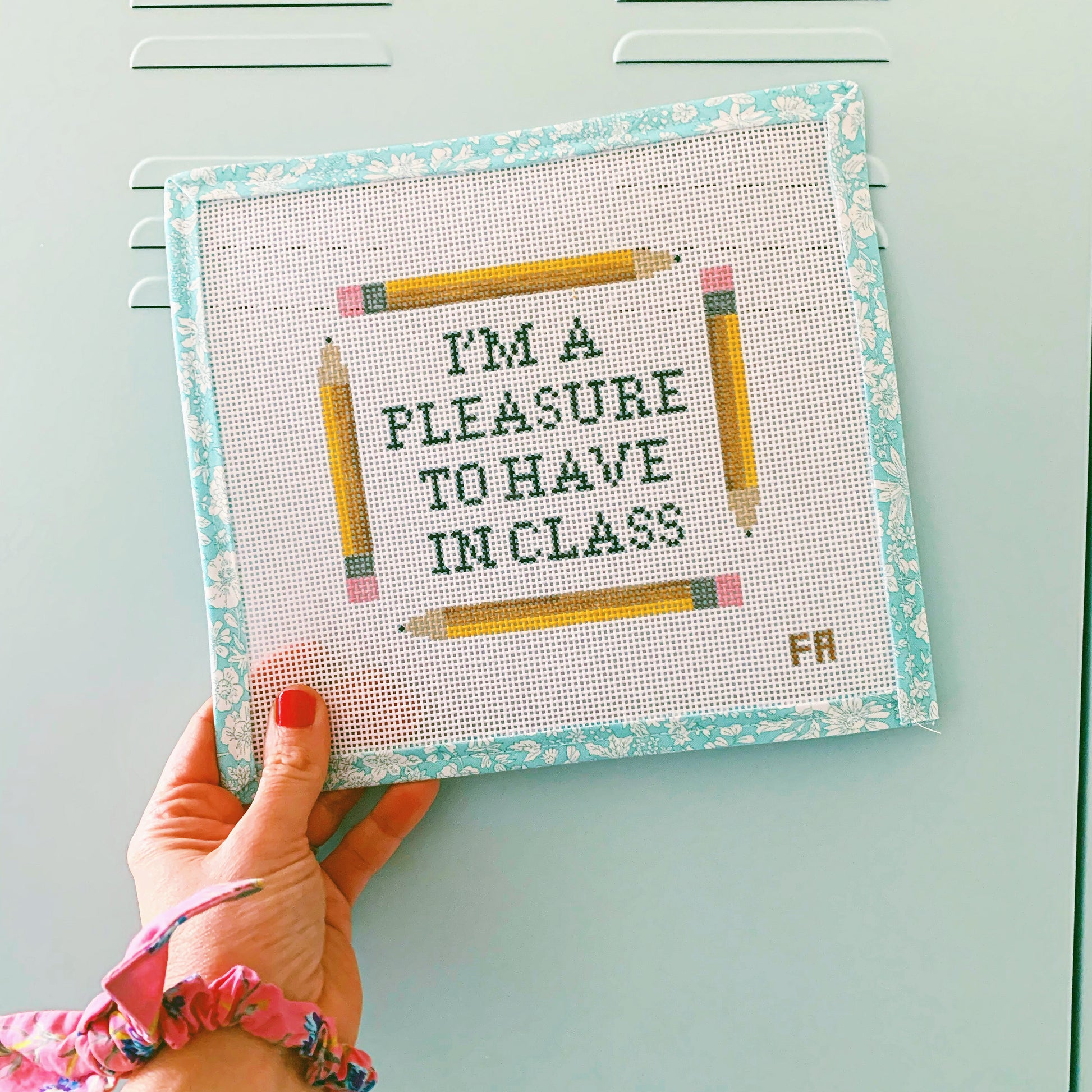 Hand holding a needlepoint canvas—printed green block letters reading “I’m a pleasure to have in class,” framed by four yellow pencils on a natural 13-mesh canvas with a turquoise floral painter’s tape border.
