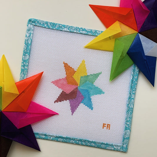 Flat-lay of Paper Star needlepoint canvas kit—vibrant eight-pointed star in red, orange, yellow, green, blue, purple, and pink printed on natural 13-mesh canvas, edged with turquoise floral painter’s tape and styled alongside folded origami stars