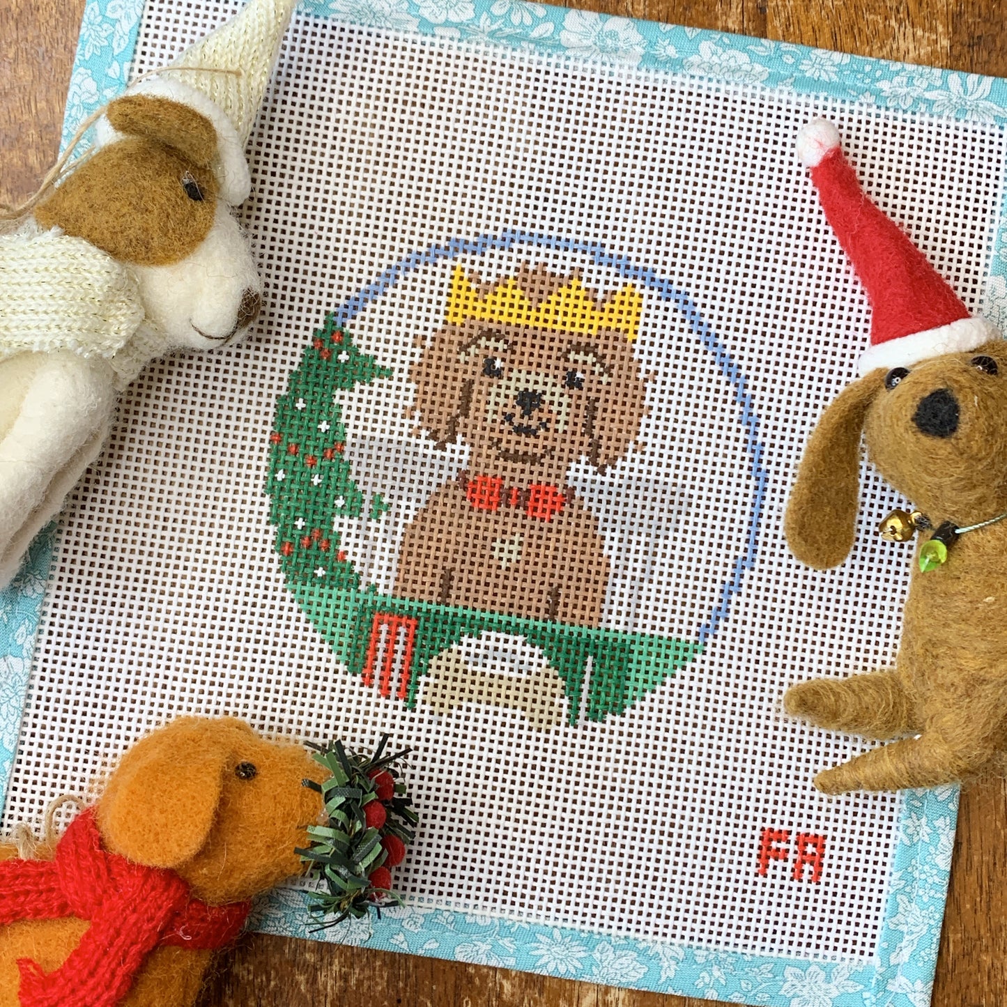 Flat-lay of a 4″ round 14 hpi needlepoint canvas brown dog with yellow crown at Christmas table with Christmas tree