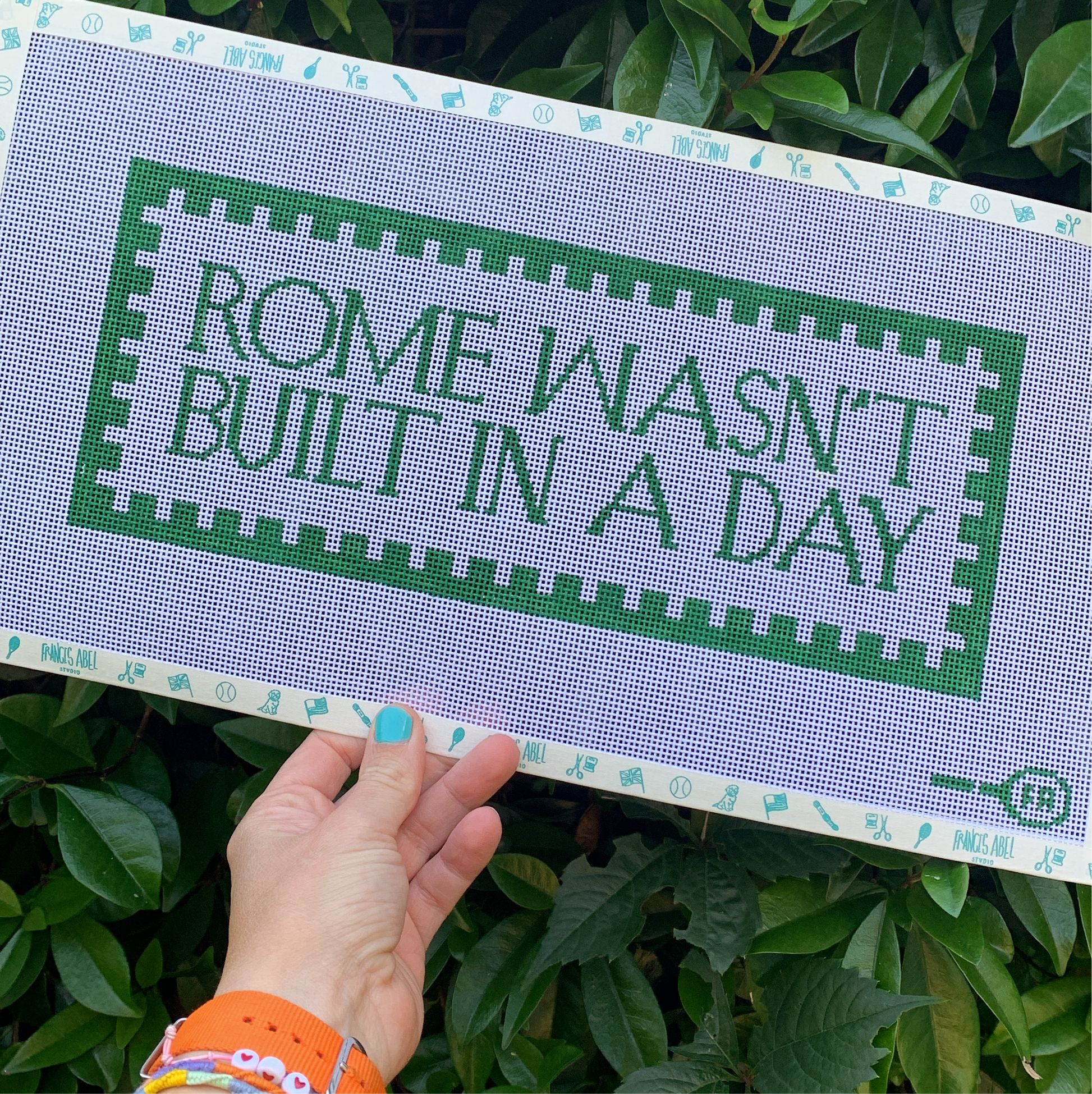 ’Rome Wasn’t Built in a Day’ needlepoint canvas kit—green block-letter text surrounded by a green checkerboard-style border on natural 13-mesh canvas