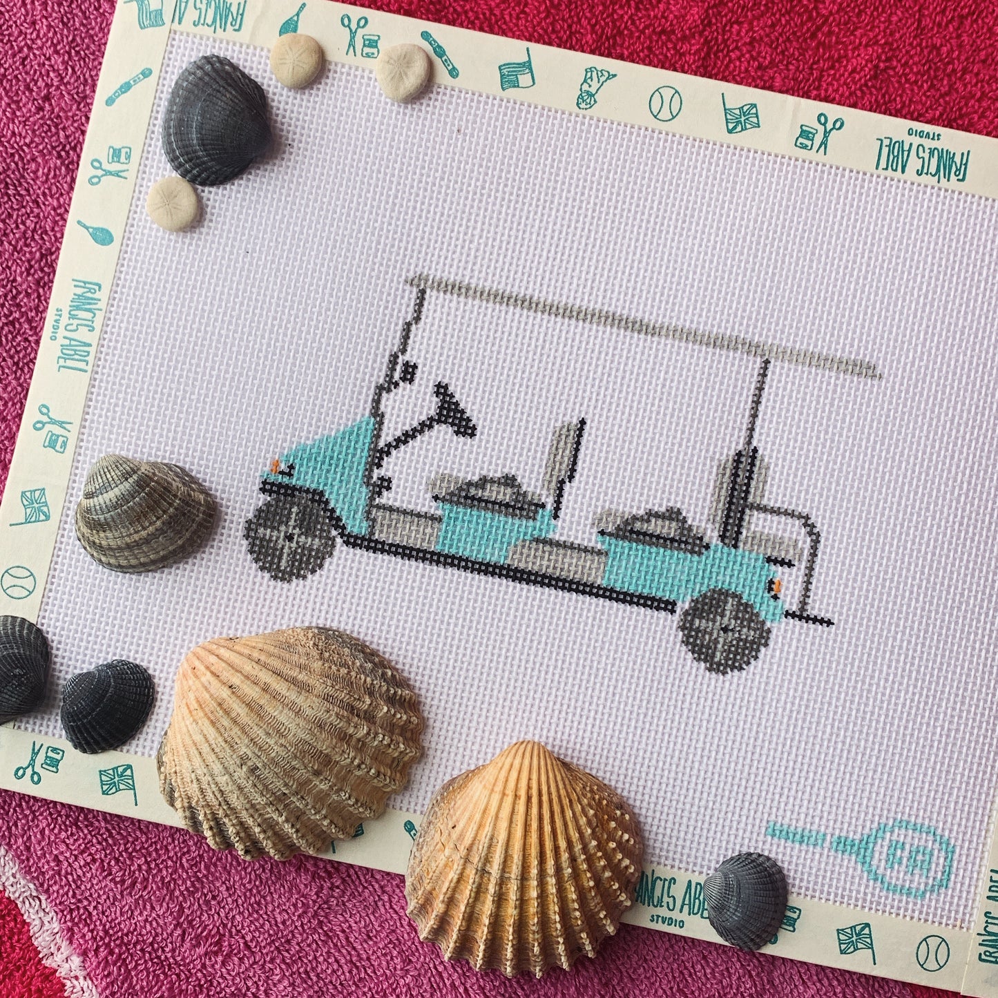 Six-seat beach cart needlepoint canvas kit—turquoise bench seats on a black metal frame and tires printed on natural 13-mesh canvas, styled flat-lay with seashells and Frances Abel border

