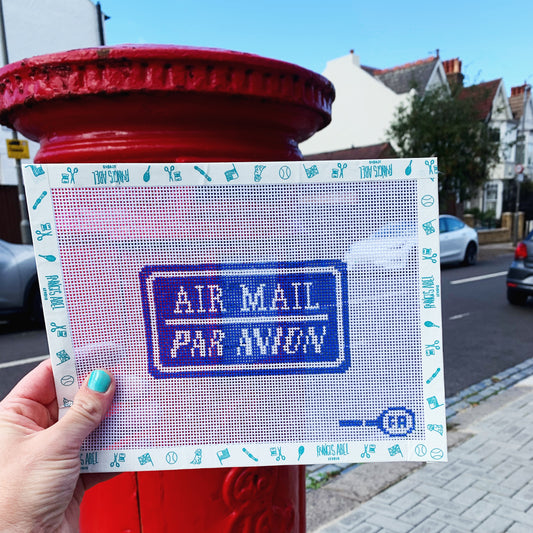 Hand holding ‘Air Mail / Par Avion’ needlepoint canvas kit—blue postal-stamp motif on natural 14-mesh 
