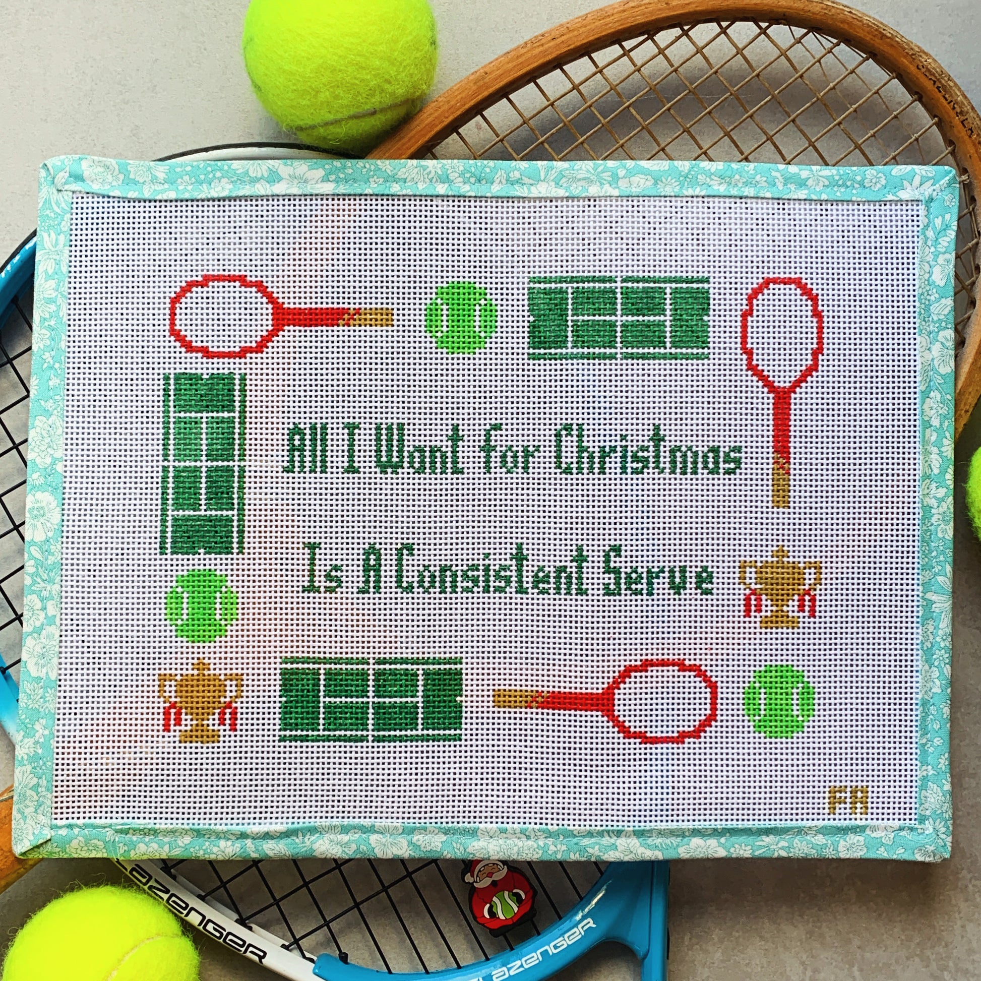 "All I Want for Christmas Is a Consistent Serve" needlepoint canvas—green block lettering framed by red tennis rackets, green tennis balls, mini tennis court grids, and tiny trophy icons on natural 14-HPI canvas, 9 × 6½″ DIY holiday tennis décor project