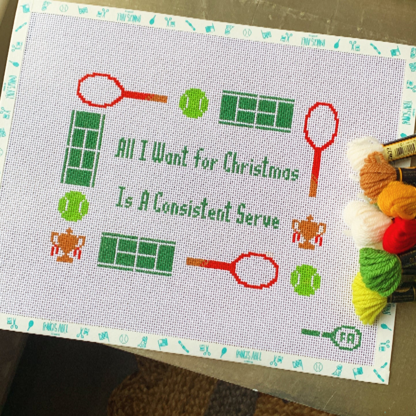 "All I Want for Christmas Is a Consistent Serve" needlepoint canvas—green block lettering framed by red tennis rackets, green tennis balls, mini tennis court grids, and tiny trophy icons on natural 14-HPI canvas, 9 × 6½″ DIY holiday tennis décor project