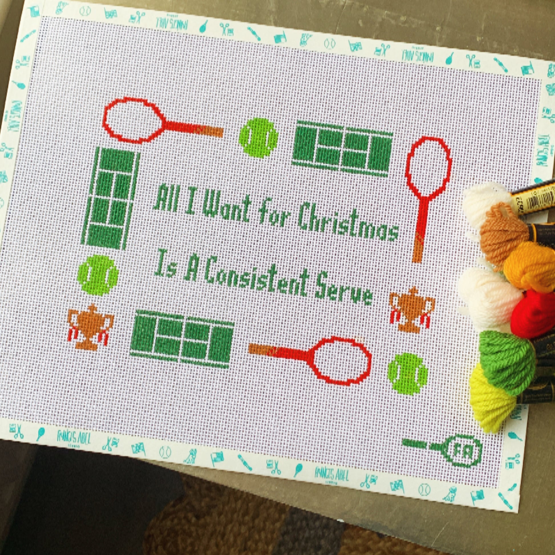 "All I Want for Christmas Is a Consistent Serve" needlepoint canvas—green block lettering framed by red tennis rackets, green tennis balls, mini tennis court grids, and tiny trophy icons on natural 14-HPI canvas, 9 × 6½″ DIY holiday tennis décor project