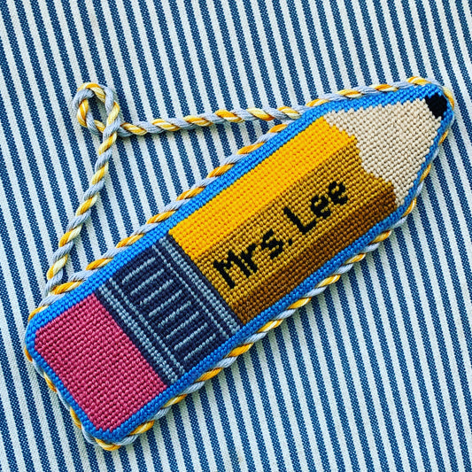 Finished custom No. 2 pencil needlepoint ornament with "Mrs. Lee" stitched in black on yellow, featuring a pink eraser, blue border, and yellow-and-gray twisted cord, displayed on blue and white striped fabric.