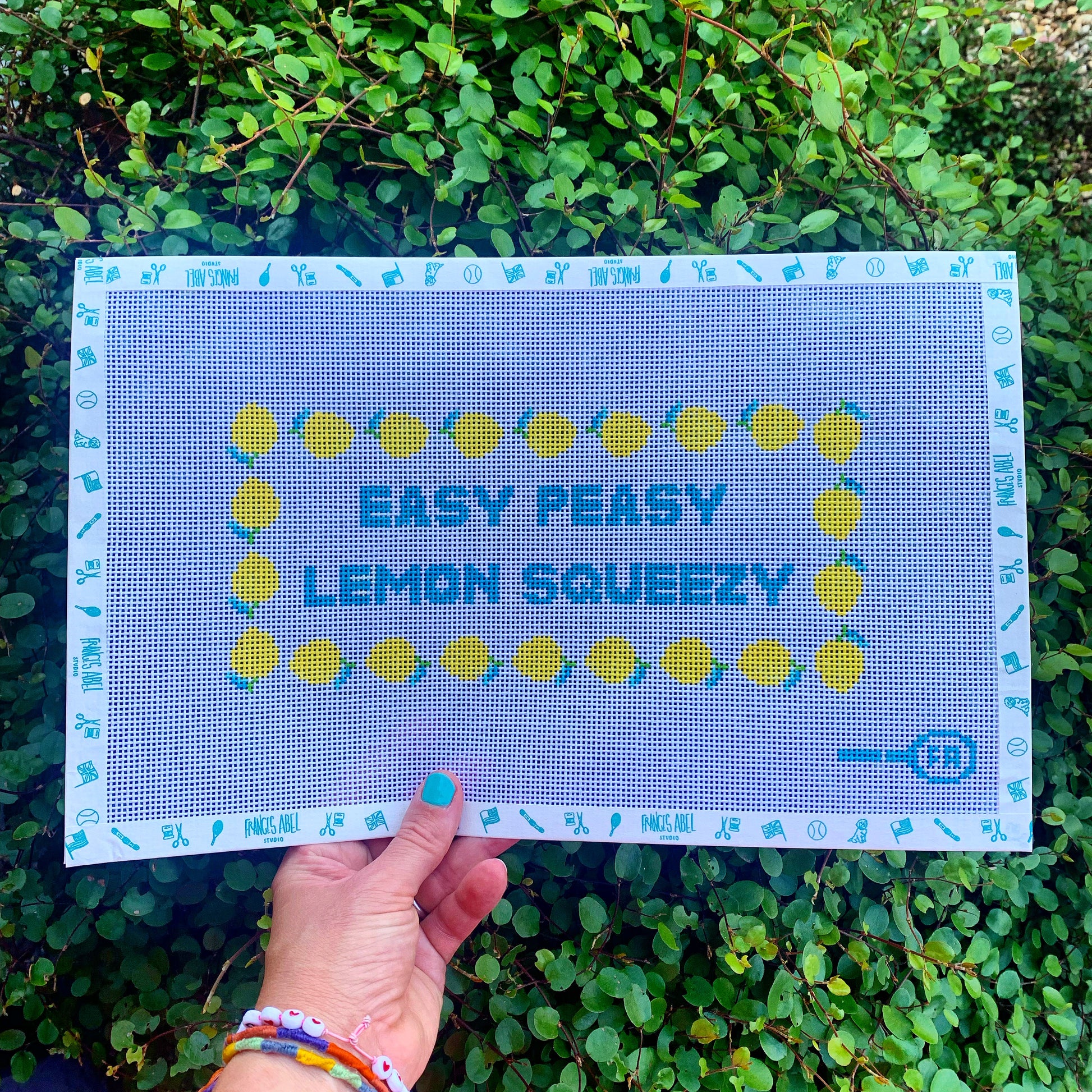 Hand holding a needlepoint canvas sheet—turquoise border of tool icons, centered teal block text “EASY PEASY LEMON SQUEEZY” framed by a ring of yellow lemon motifs on natural 13-mesh