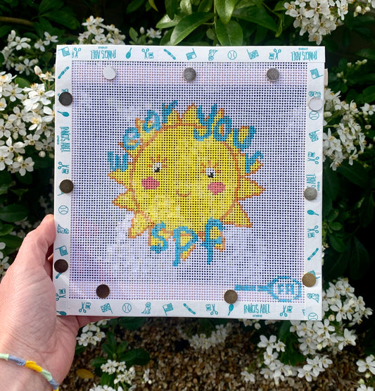 Friendly Sun “Wear Your SPF” needlepoint canvas kit—smiling yellow sun wearing sunglasses with bold “Wear Your SPF” text on natural 13-mesh canvas, 4½ × 4½″ DIY summer décor project
