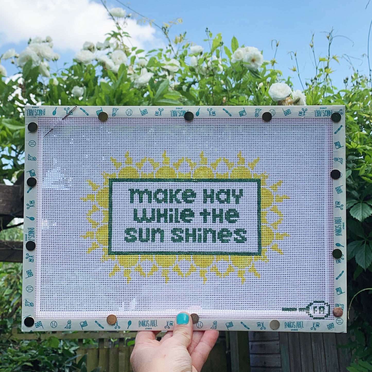 ‘Make Hay While the Sun Shines’ needlepoint canvas kit—green block-letter text in a rectangular frame, surrounded by repeating yellow sunburst motifs on natural 13-mesh canvas 