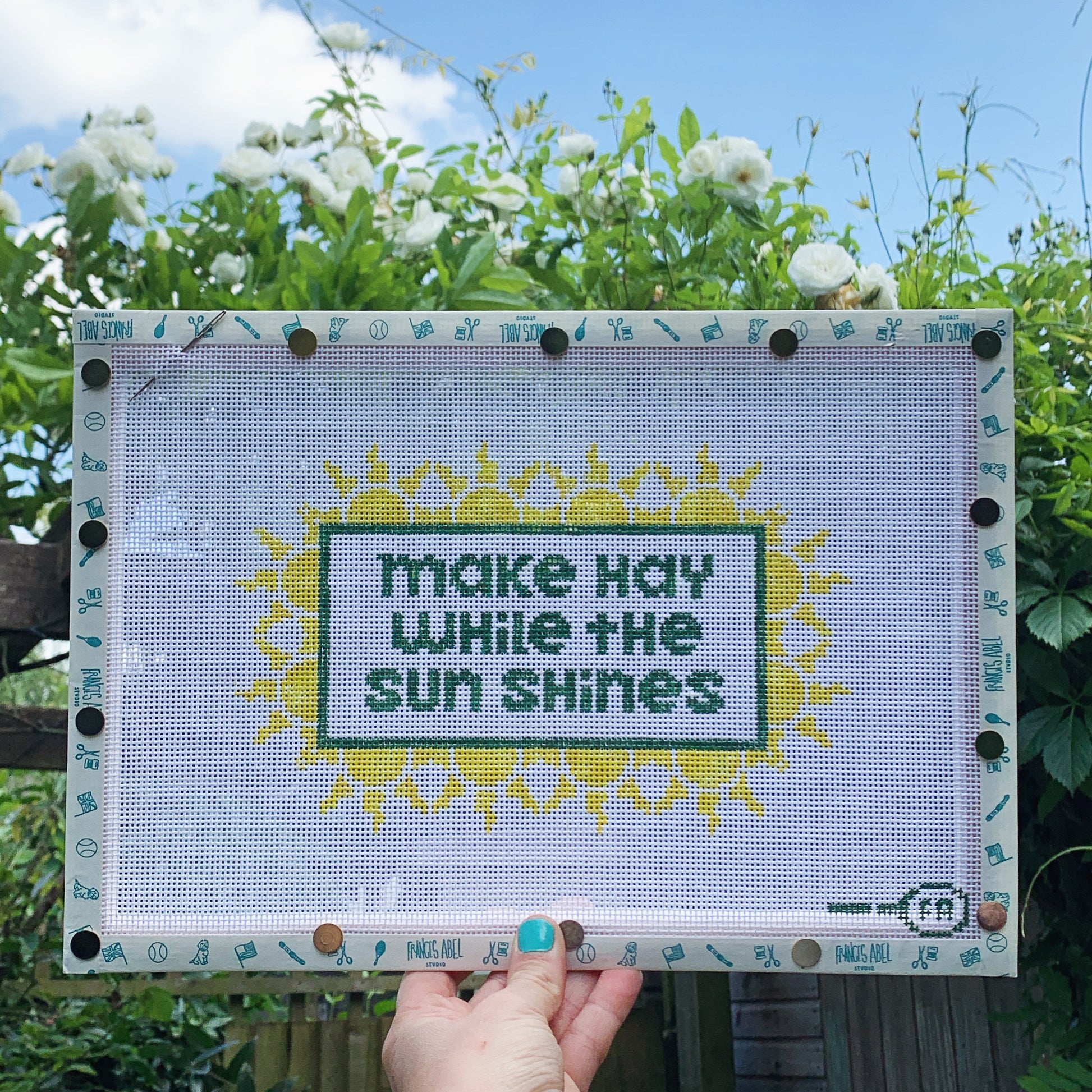 ‘Make Hay While the Sun Shines’ needlepoint canvas kit—green block-letter text in a rectangular frame, surrounded by repeating yellow sunburst motifs on natural 13-mesh canvas 