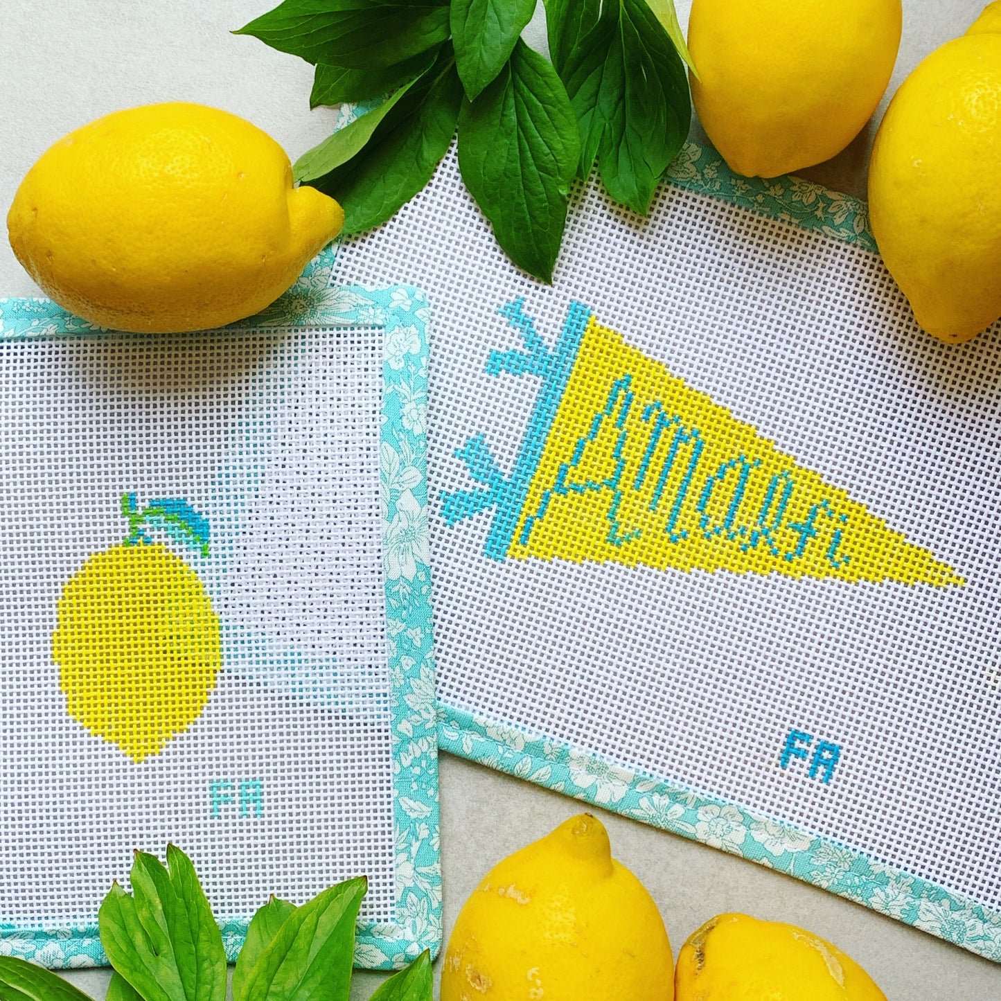 Mini lemon and Amalfi pennant needlepoint canvases with turquoise floral trim, surrounded by fresh lemons and green leaves on a light surface.