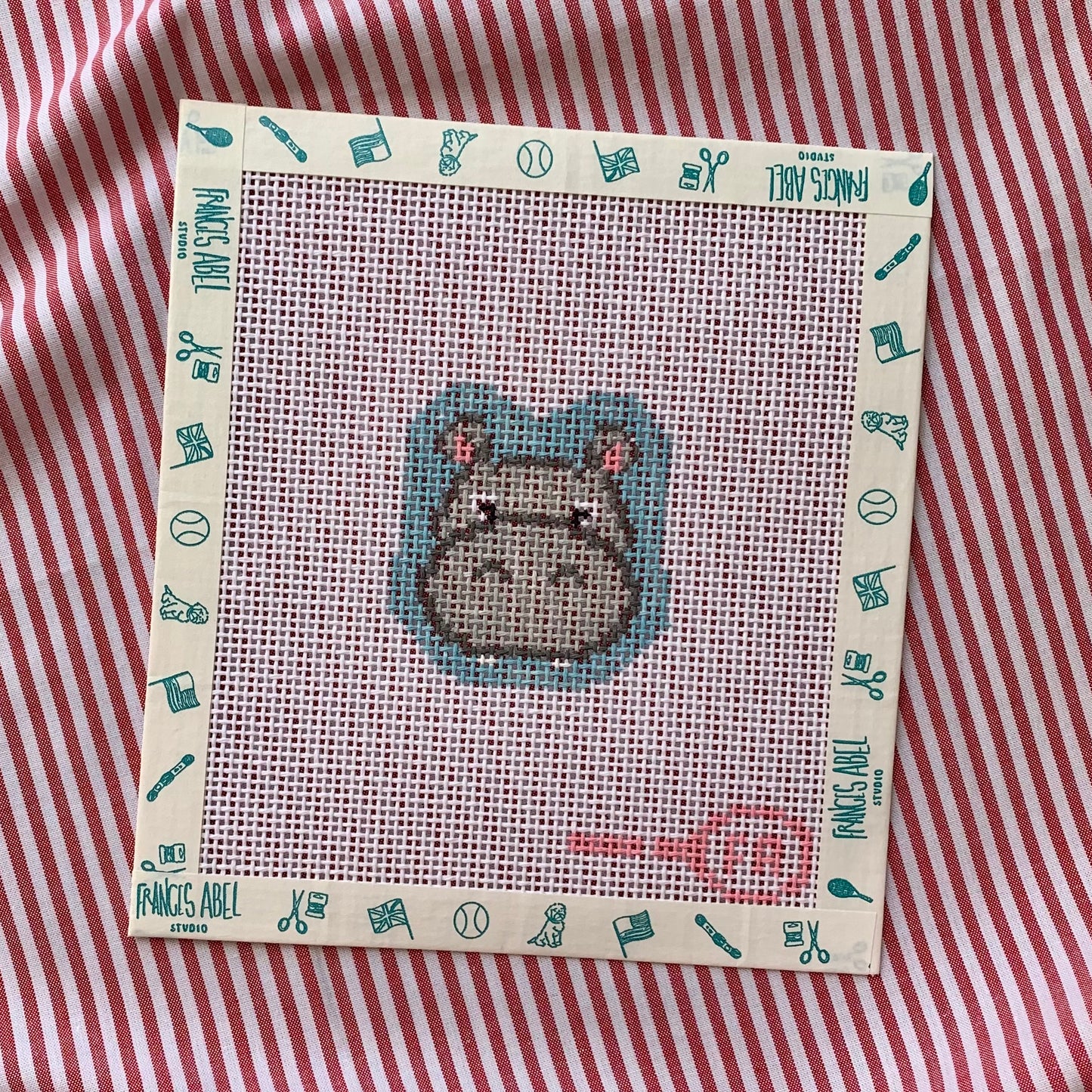 Mini Miss Hippo needlepoint canvas kit—grey hippo head with pink ear accents at center of natural 13-mesh canvas, surrounded by teal icon-patterned frame, styled on red-and-white striped background 
