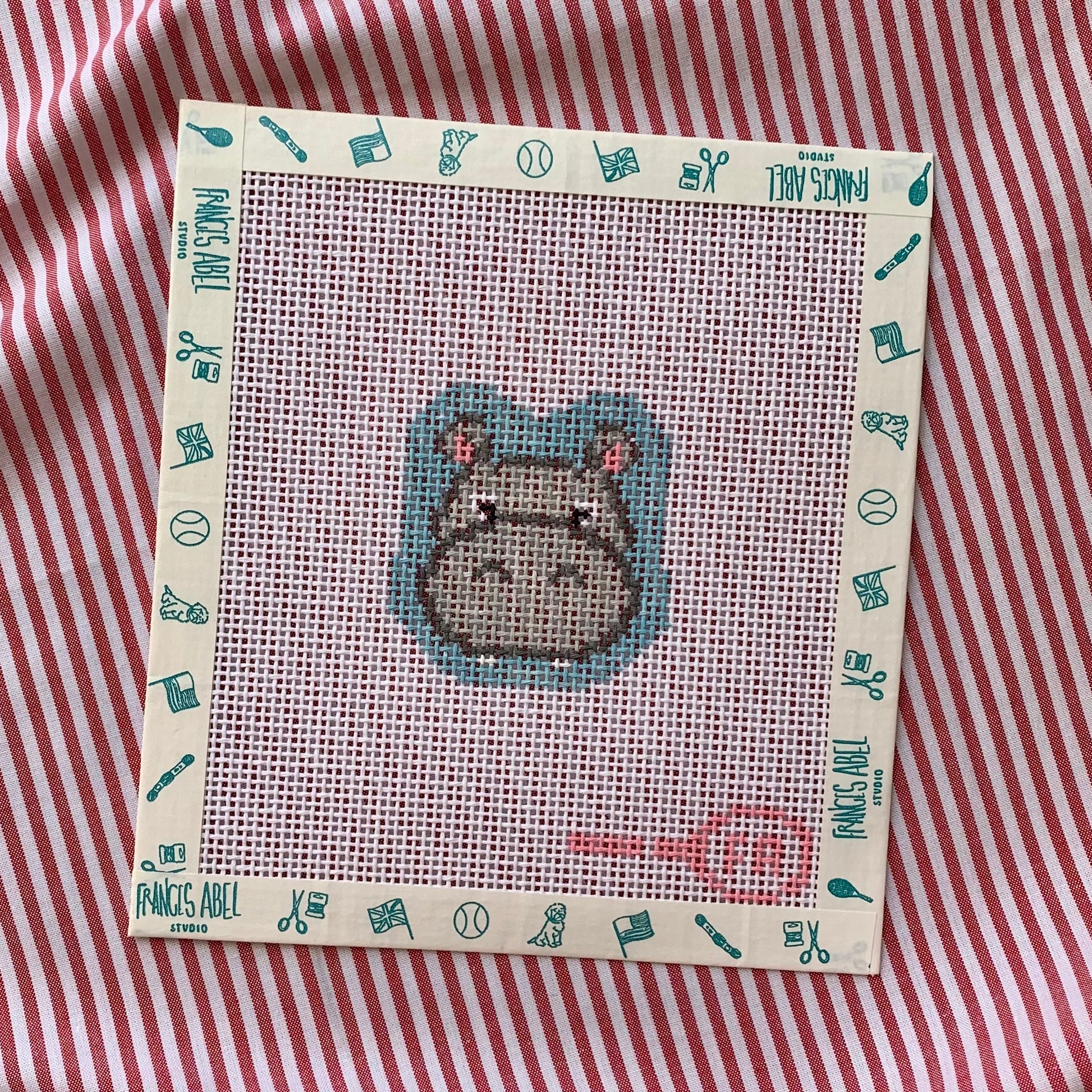 Mini Miss Hippo needlepoint canvas kit—grey hippo head with pink ear accents at center of natural 13-mesh canvas, surrounded by teal icon-patterned frame, styled on red-and-white striped background 
