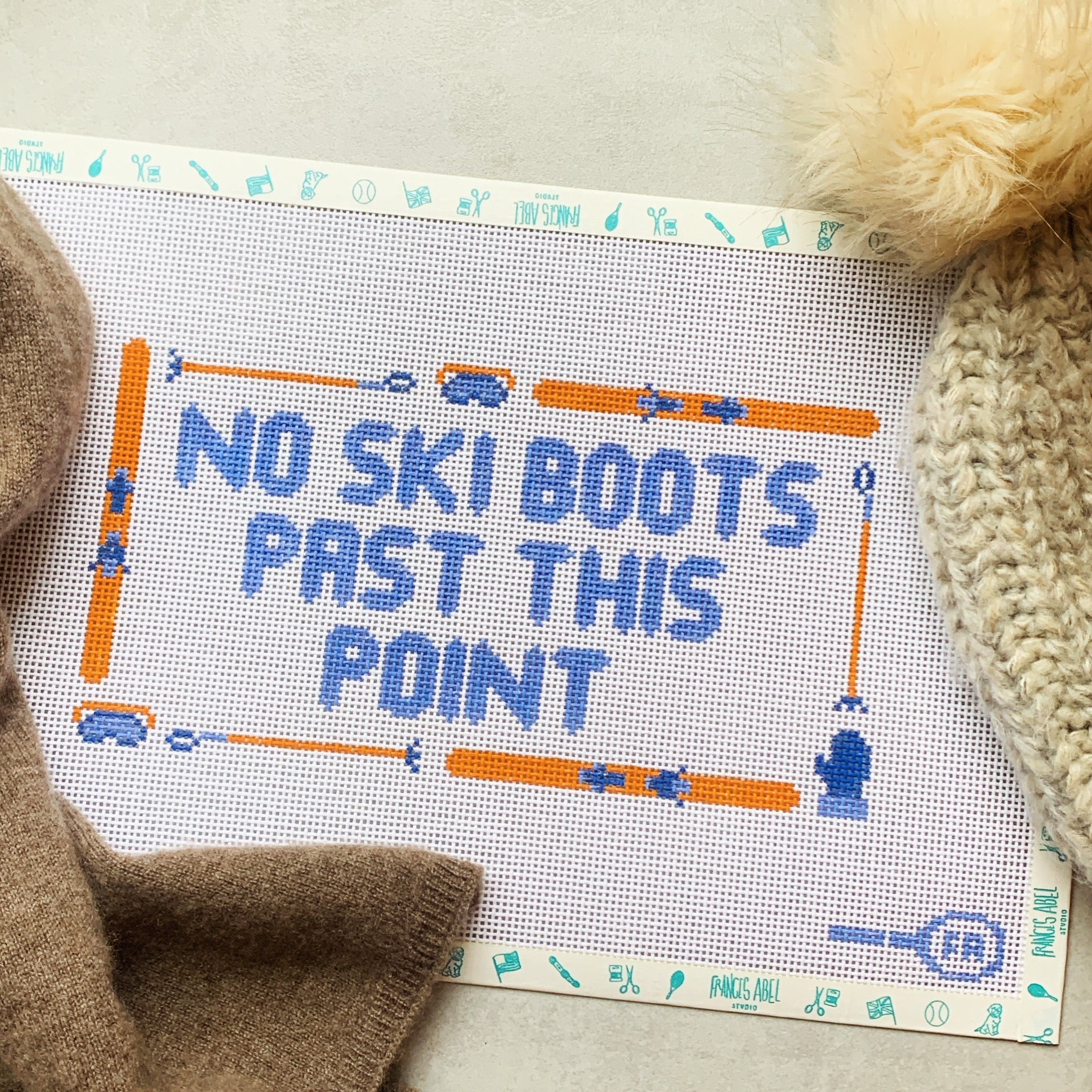 No Ski Boots Past This Point needlepoint canvas kit—bold blue block lettering framed by orange skis, poles, and ski-boot icons on white 13-mesh canvas, styled with a cream pom-pom hat and brown scarf