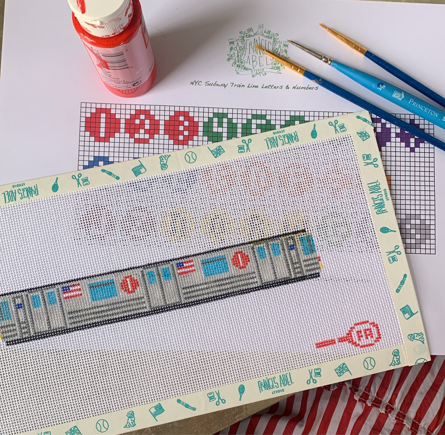 Partial NYC Subway Key Fob needlepoint canvas taped to a board—showing a stitched gray subway car with blue windows and red line logos, laid over its printed chart, with a red paint jar and three paintbrushes resting on a white surface 
