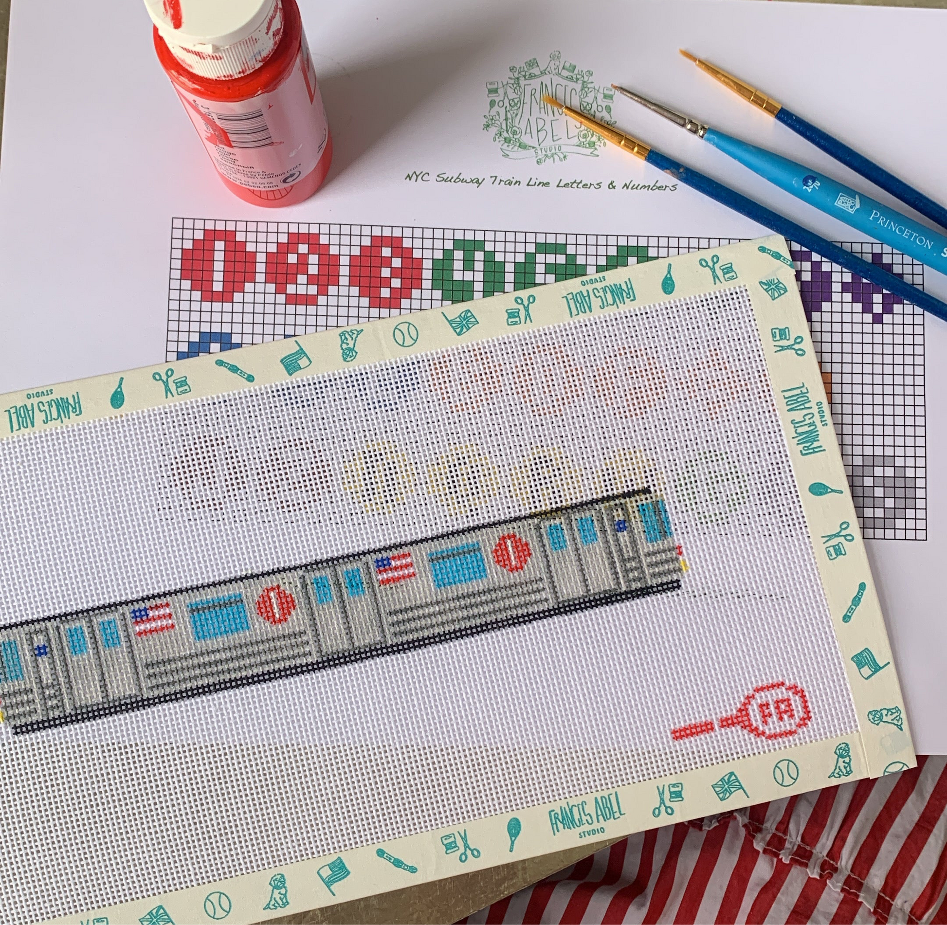 Partial NYC Subway Key Fob needlepoint canvas taped to a board—showing a stitched gray subway car with blue windows and red line logos, laid over its printed chart, with a red paint jar and three paintbrushes resting on a white surface 
