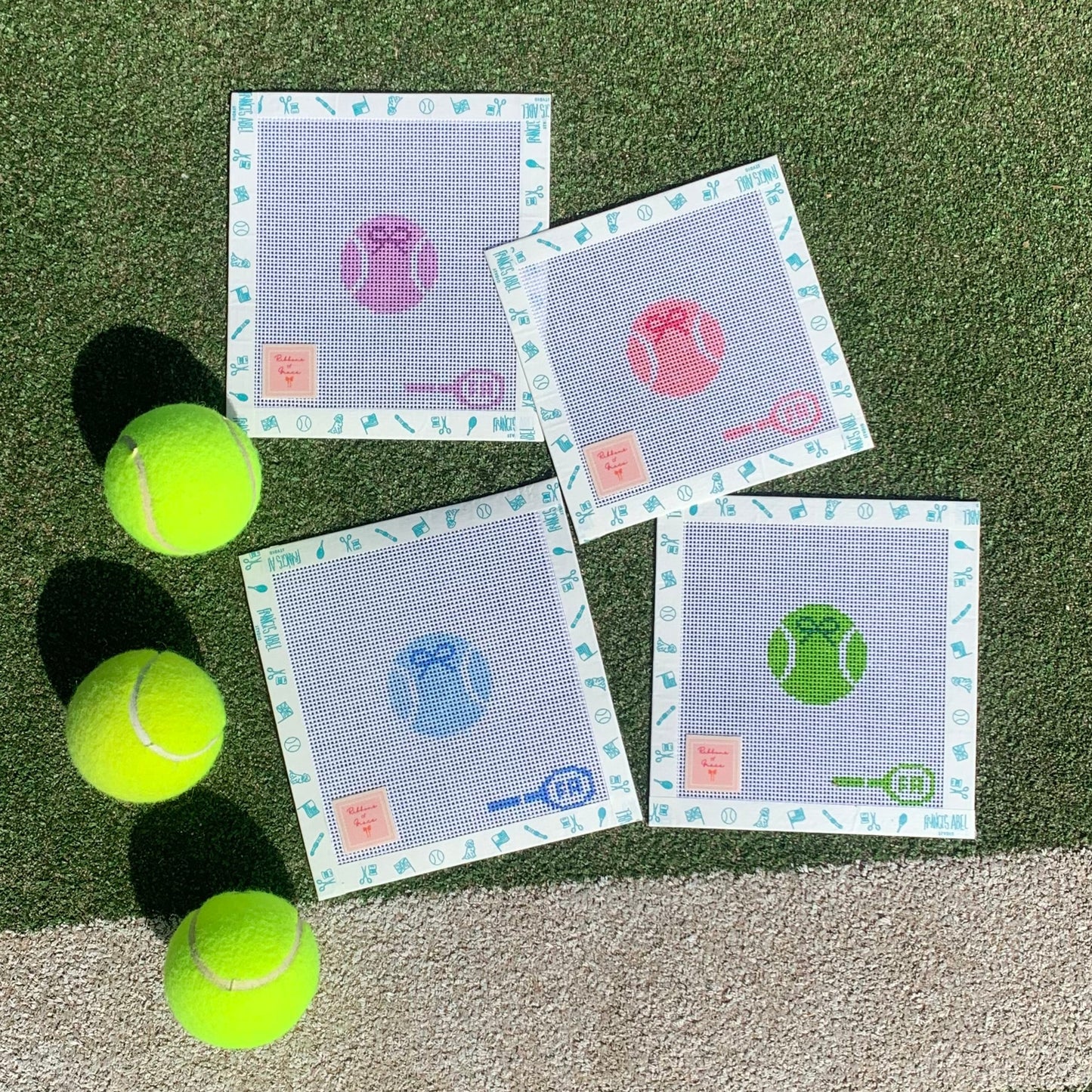Four square needlepoint canvases—custom-painted tennis ball designs in purple, pink, blue, and green on 14-HPI mesh, arranged on artificial turf alongside three neon-yellow tennis balls 