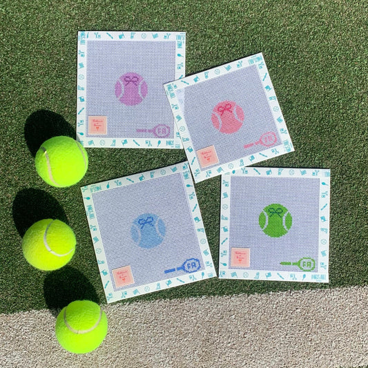Four square needlepoint canvases—custom-painted tennis ball designs in purple, pink, blue, and green on 14-HPI mesh, arranged on artificial turf alongside three neon-yellow tennis balls 