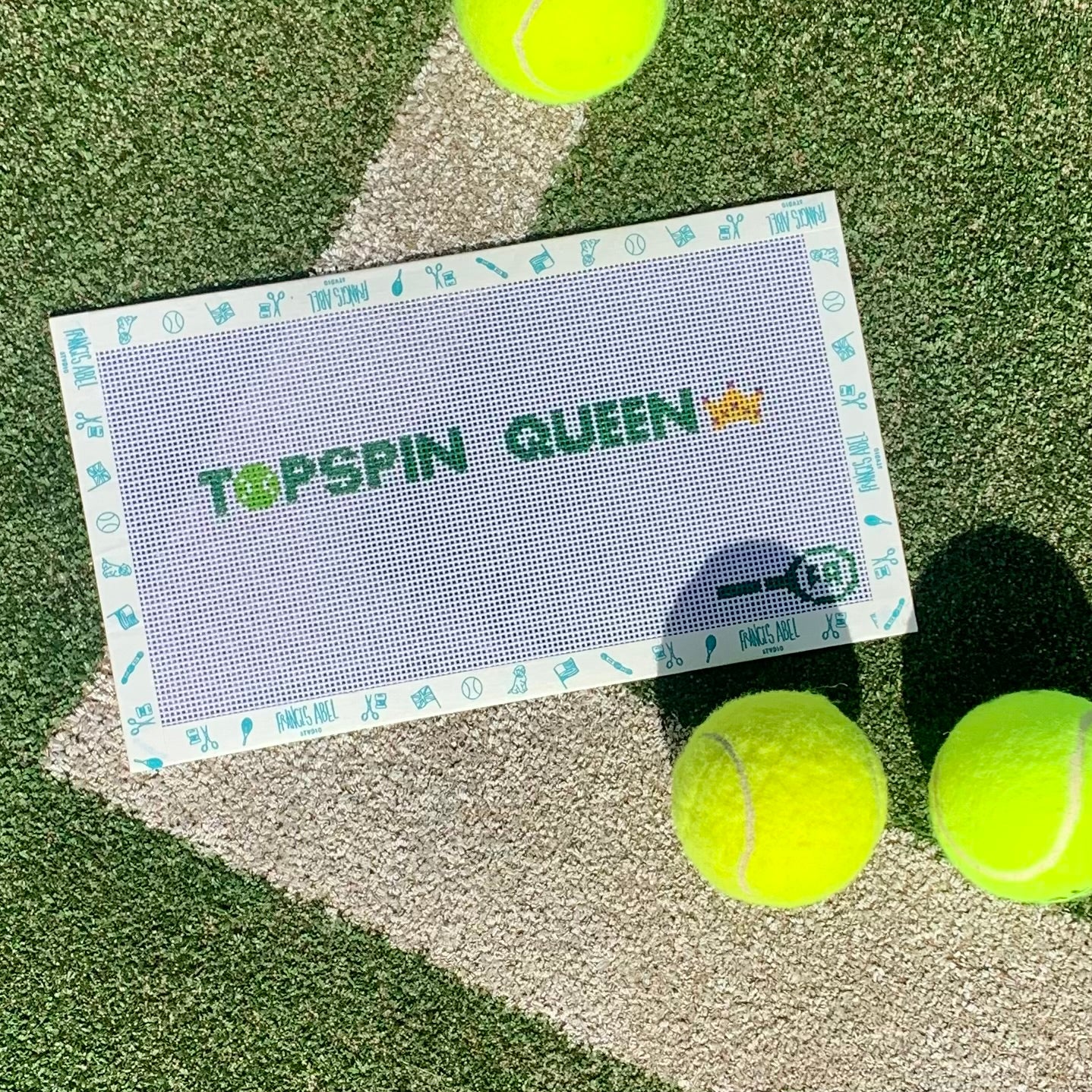 Flat-lay of Topspin Queen key-fob needlepoint canvas on tennis turf—3/4 × 7½″ 14 hpi canvas printed with green block letters “TOPSPIN QUEEN,” a yellow tennis-ball “O,” and a key-fob outline, styled with neon tennis balls